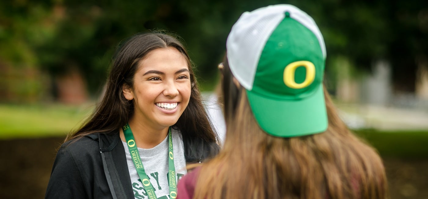 One student with dark hair and a UO green and yellow lanyard smiling at another student wearing a UO baseball cap.
